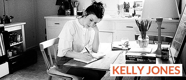 Kelly Jones writing a diary at a kitchen table near a stove a plant, french vocabulary book, chair and lamp.